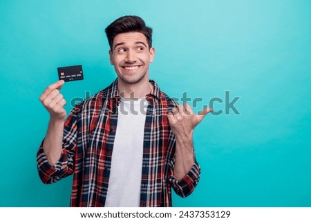 Photo of cheerful dreamy man dressed plaid shirt rising bank card pointing thumb empty space isolated turquoise blue color background