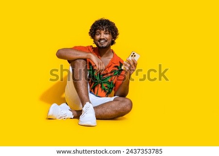 Full size photo of optimistic handsome man dressed print shirt holding smartphone in palm sit on floor isolated on yellow color background