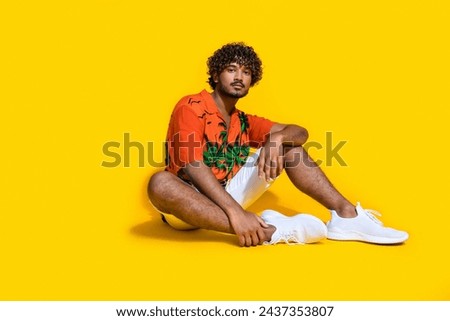 Full size photo of confident serious man dressed print shirt white shorts shoes sitting on floor isolated on yellow color background