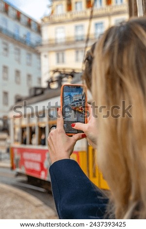 Photograph taken from the back of a woman leaning out of a tram trolley window in Lisbon Portugal taking a photograph with her cell phone she has long blond hair and red nail polish