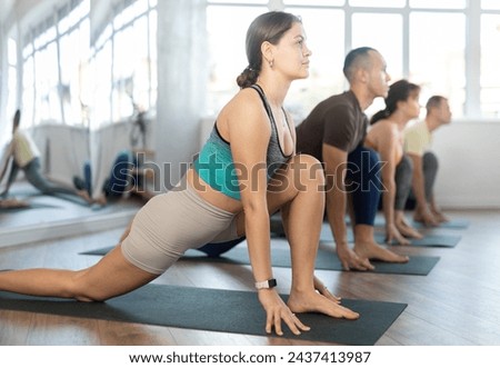 Group of young athletes practice hatha yoga in studio during class. Lovers of active lifestyle on yoga mat perform doing version of warrior pose virabhadrasana in gym Royalty-Free Stock Photo #2437413987
