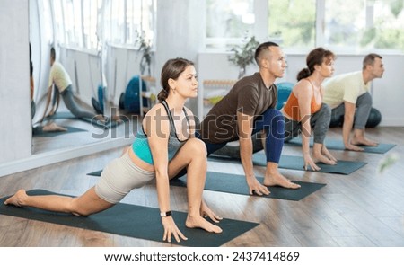 Group of young athletes practice hatha yoga in studio during class. Lovers of active lifestyle on yoga mat perform doing version of warrior pose virabhadrasana in gym Royalty-Free Stock Photo #2437414869