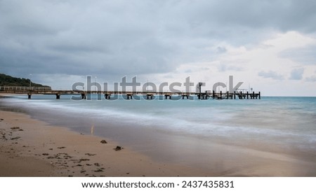 Dawn on an overcast day with a view of a fishing and small boat jetty on a tropical beach with coastal rainforest and smooth sea because of long exposure photography at Mission Beach in Australia.