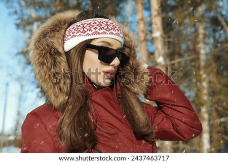 Winter holidays. Winter fashion. Beautiful fashion model girl posing in a red down jumpsuit and sunglasses in a winter snowy park.