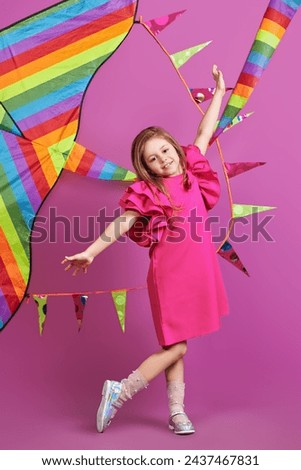 Summer holidays. Happy cute girl in a bright fuchsia dress posing cheerfully against a rainbow kite and pink background. Children's fashion. Dreams and imagination.