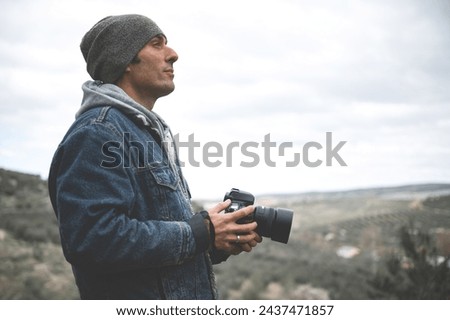 Confident side portrait of attractive young man, a travel photographer holding professional digital camera, capturing beautiful view while hiking in mountains in the early spring. Cold weather outside