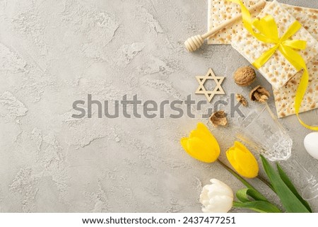 Chic Pesach setting from a top view, with matzah in a ribbon, glass, walnuts, egg, honey dipper, and tulips, arranged on a minimalist grey concrete background, with placeholder for text