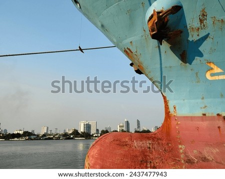 Bow side with Anchor on large cargo ship's anchor being pulled. Blue and red ship, While docked at the pier by large ropes on the river, in the background is a view of the city and transport concept