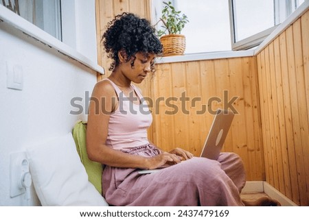 A young woman in casual attire focuses intently on her laptop in a cozy, well-lit wooden alcove. The relaxed home setting emphasizes comfort and productivity. Royalty-Free Stock Photo #2437479169