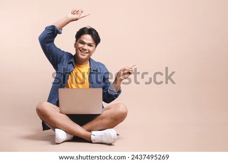 Full length of young Asian man sitting on floor with laptop on his laps, pointing aside to the empty space for ads 