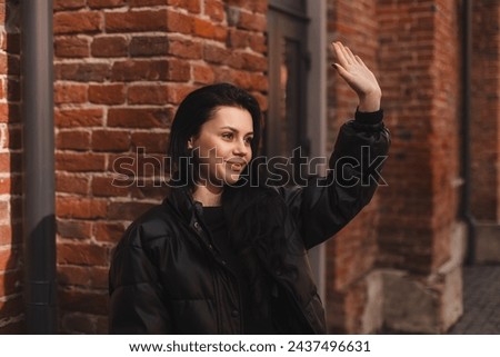 Happy attractive woman in black puffer jacket, walk outdoors, meeting her friends or love, showing high five hey hello hi sign. She is joyful to meet her acquainted by chance. Girl wave her hand.
