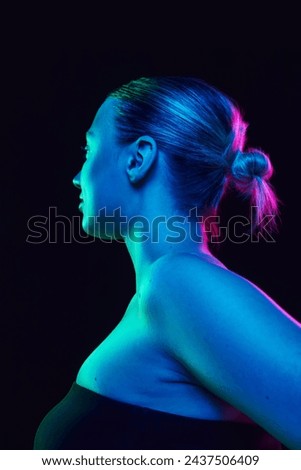 Profile photo of young woman with bare shoulders with bun hairstyle looking away in neon light against black studio background. Concept of spa procedures, cosmetology, skincare products. Ad