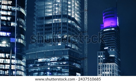 Skyscraper at construction process by night. Modern high-rise building under construction next to glass skyscrapers by night.