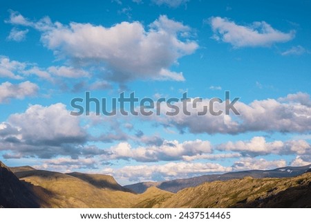 Mountain range and hills illuminated by sunset light. Rocks in gold sunrise colors. Vivid golden rockies in sunlight under clouds of sunset tones in blue sky. Shadows of clouds on rocky mountain ridge