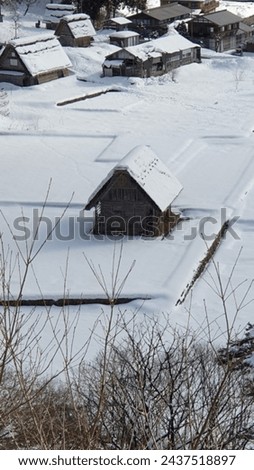 a small solitary traditional house in the middle of snow field with a roof covered with snow. At the back ground are several similar houses. Royalty-Free Stock Photo #2437518897