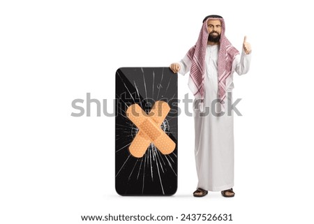 Saudi arab man in ethnic clothes gesturing thumbs up and standing next to a mobile phone with cracked screen isolated on white background