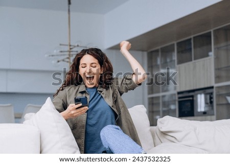 A woman sits on a sofa, her arm raised triumphantly as she looks at her smartphone, embodying a moment of victory or good news received. Her exuberant expression conveys pure joy. Exuberant Woman Royalty-Free Stock Photo #2437532325