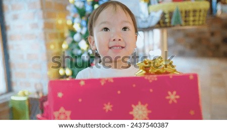 A young child with wide eyes holding a large red gift box and smile while standing in front of a vibrant Christmas tree, capturing the wonder of holiday celebrations.
