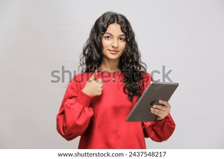 Attractive Pakistani smiling girl in red sweatshirt shirt using tablet showing thumbs up. Woman with tablet pc, isolated on white background
