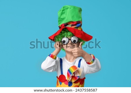 Cute little boy in clown costume with funny glasses showing thumb-up on blue background. April Fools Day celebration Royalty-Free Stock Photo #2437558587