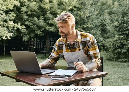 hard working handsome man with beard using laptop and clipboard to analyze resources on farm Royalty-Free Stock Photo #2437565687