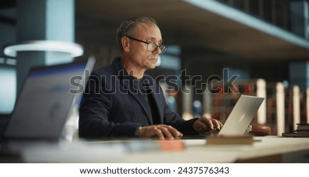 Knowledgeable Teacher Using Laptop Computer in a Library. Smart Middle Aged Male Educator Preparing Lectures for College Students, Checking Exams, Browsing and Researching Internet Articles