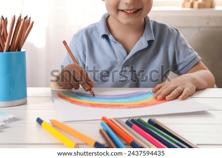 Little boy drawing rainbow with pencil at white wooden table indoors, closeup. Child`s art