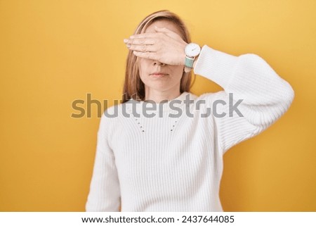 Young caucasian woman wearing white sweater over yellow background covering eyes with hand, looking serious and sad. sightless, hiding and rejection concept  Royalty-Free Stock Photo #2437644085