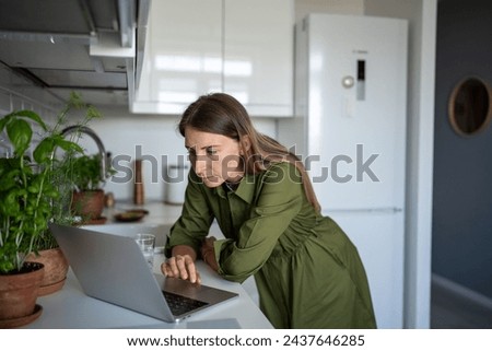 Tired concentrated woman freelancer trying to work at home kitchen standing with laptop after long sitting time at desk. Serious focused female looking on screen computer searching info, scrolling web