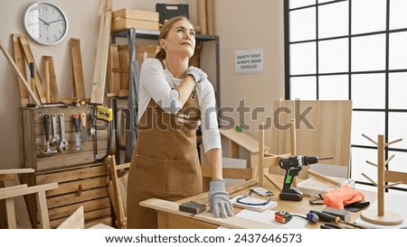 Mature woman in apron feeling shoulder pain while working in a carpentry workshop with tools around.