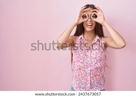Young hispanic woman with long hair standing over pink background doing ok gesture like binoculars sticking tongue out, eyes looking through fingers. crazy expression. 