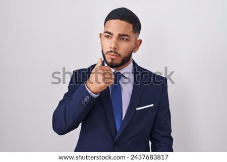 Young hispanic man wearing business suit and tie with hand on chin thinking about question, pensive expression. smiling with thoughtful face. doubt concept.  Royalty-Free Stock Photo #2437683817
