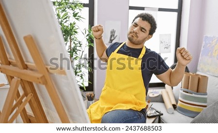 A young bearded man stretches his back in a colorful art studio while wearing a yellow apron.