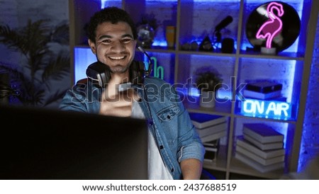 A cheerful man with headphones gesture towards camera in a neon-lit gaming room at night.
