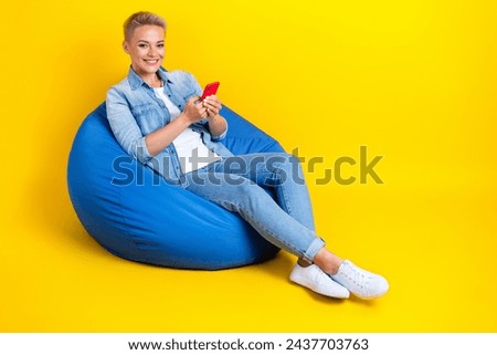 Full size photo of pretty young girl sit bean bag hold device influencer dressed stylish denim outfit isolated on yellow color background