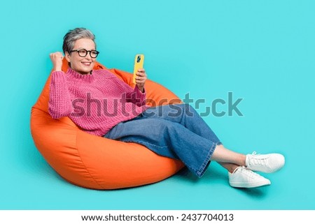 Full size photo of satisfied person dressed knitwear jumper sit on pouf look at smartphone win bet isolated on turquoise color background