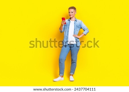 Full size photo of pretty young girl hold telephone read message dressed stylish denim outfit isolated on yellow color background