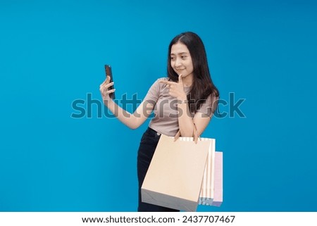 beautiful asian woman is looking at her smartphone with a smile, her other hand holding a shopping bag isolated on blue background