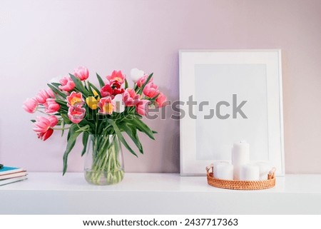 Modern interior design with tulip flowers bouquet in vase, white picture frame mockup, candles on white console on pink wall background. Poster design mockup. Minimalist Home decor. Selective focus.