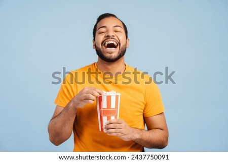 Overjoyed african american man holding bucket with popcorn, eating snacks, laughing, watching movie isolated on blue background. Cinema, advertisement concept 
