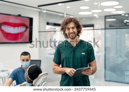 Proud young dental surgeon wearing green medical uniform holding digital tablet standing in his clinic with patient in dentist's chair and nurse in background.