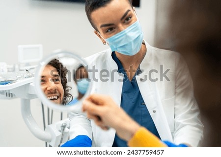 Happy male patient looking at mirror after dental treatment In a clinic. Cheerful man enjoying his beautiful smile. Female orthodontist communicating with satisfied patient after successful procedure.
