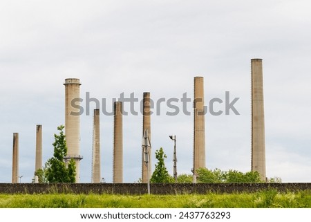 A row of tall, disused industrial smokestacks stands against a cloudy sky, remnants of a bygone manufacturing era, showcased here in a quiet state of neglect. Royalty-Free Stock Photo #2437763293