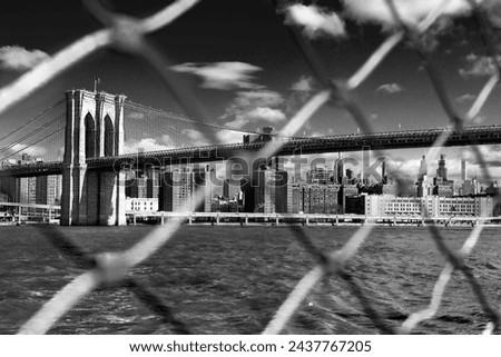 black and white new york city - the Brooklyn Bridge framed through the links of a chain cyclone fence with the East River and midtown Manhattan buildings