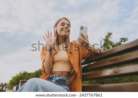 Young woman wears orange shirt casual clothes use mobile cell phone listen to music in earphones wave hand sit on bench walk rest relax in spring city park outdoors on nature. Urban lifestyle concept