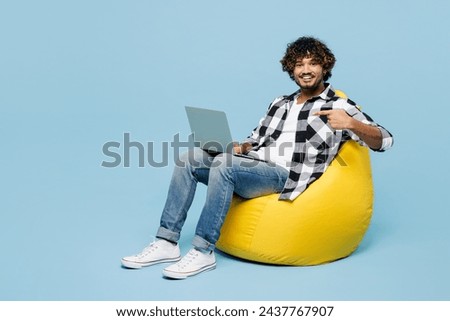 Full body young Indian IT man he wears shirt white t-shirt casual clothes sit in bag chair hold use work point on laptop pc computer isolated on plain pastel light blue background. Lifestyle concept