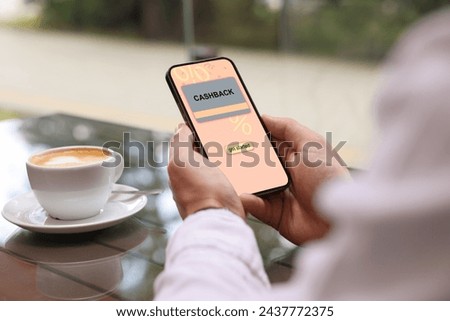 Cashback. Man holding smartphone at table indoors, closeup. Illustration of credit card and percent signs on device screen