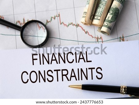 FINANCIAL CONSTRAINTS - words on a white sheet against the background of a chart, magnifying glass and banknotes Royalty-Free Stock Photo #2437776653