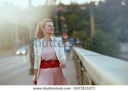 happy modern 40 years old woman in pink dress and white jacket in the city on the bridge. Royalty-Free Stock Photo #2437815371