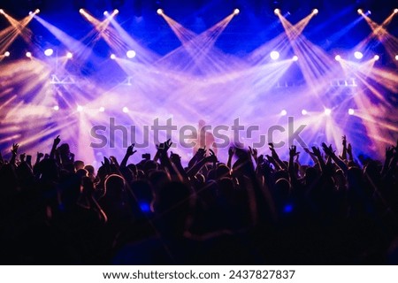 Silhouettes of people dancing and rising hands while having fun on the open air concert at night. Performer is singing on stage and making good atmosphere. There are purple and blue stage lights. Royalty-Free Stock Photo #2437827837